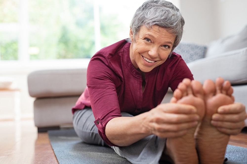 Foot Care and Aging. Closeup of senior woman stretching to touch toes while sitting on yoga mat. Portrait of mature woman doing her stretches at home while looking at camera. Happy healthy lady doing yoga exercises.