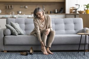 Foot Care and Aging. Female came home sit on sofa took off shoes, touch ankle doing self-massage of tired feet, suffers from varicose, painful feelings discomfort after long workday on heels, need rest, orthopedic insoles