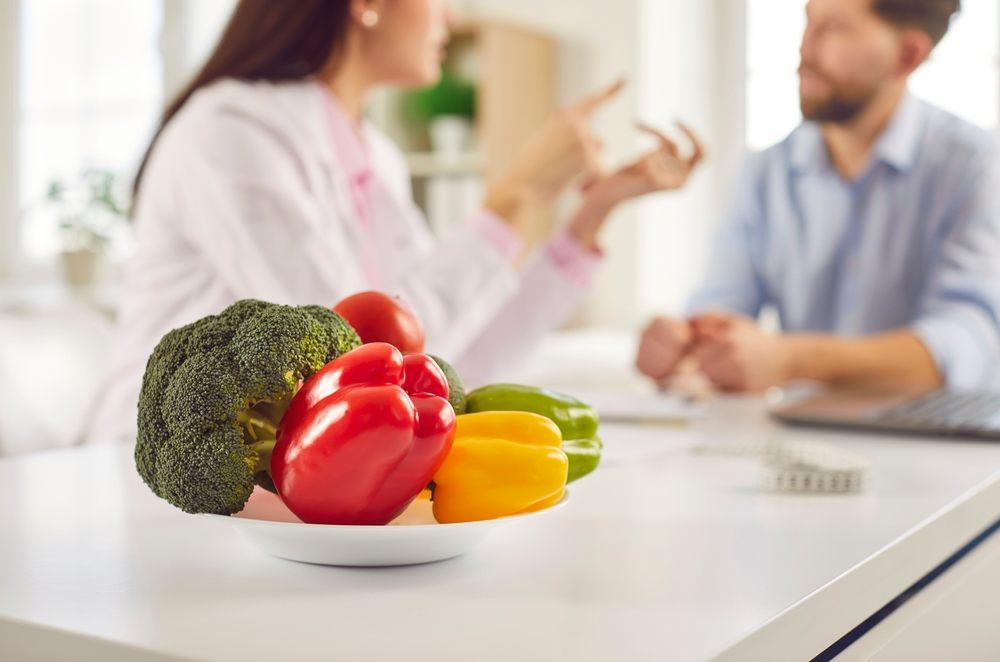 Fueling Your Ottawa Winter: Nutrition Tips for Staying Energized and Healthy? Fresh healthy vegetables on a plate in the foreground in nutritionist doctor's office. Dietician woman sitting on workplace and giving consultation to man patient. Healthy nutrition concept.