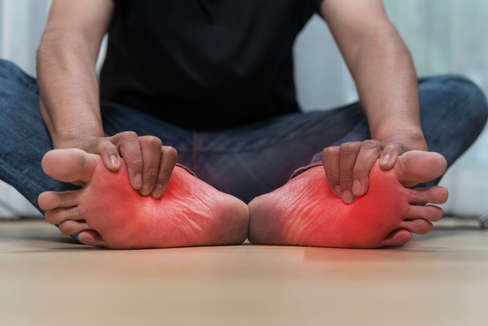 Pedorthic Solutions: Improving Foot Comfort & Function? Close up of man sitting on floor holding both feet with visible redness and pain, symptoms of inflammation, plantar fasciitis, neuropathy, gout, or diabetic foot problems related to chronic conditions.